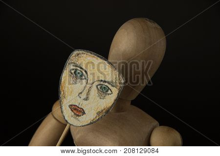 Wooden puppet doll on hinges holds a mask in hands and covers her face on a black background
