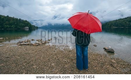 Woman with red umbrella contemplates on rain in front of a lake. Sad and lonely female person looking into distance.