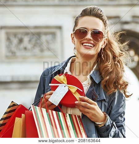 Stylish Christmas in Paris. Portrait of happy elegant woman in trench coat with shopping bags and Christmas present near Arc de Triomphe in Paris France
