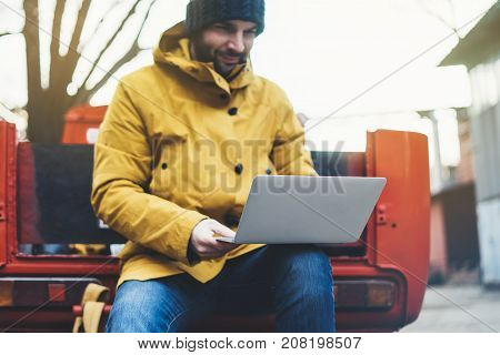 Hipster with yellow backpack jacket and cap sitting on retro red car freelance using computer open laptop in spring street outdoor tourist man typing on keyboard traveler connect internet process in workspace
