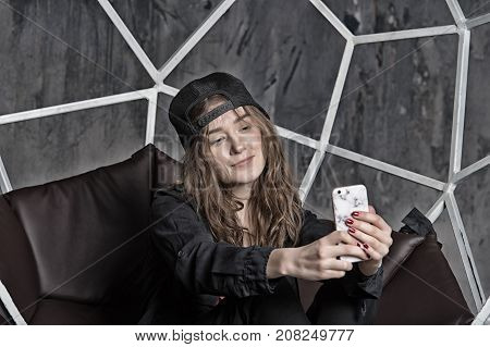 Woman or girl in baseball cap making selfie with smartphone on brown chair. Technology and mobile device. Self expression concept vintage filter