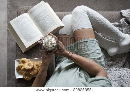pretty girl reading book at home on couch with hot chocolate milk and cookies