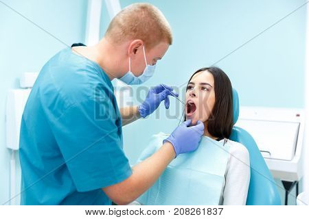 A dermatologist, a blonde man in purple gloves, examines the teeth of a young pretty black-haired girl at his reception