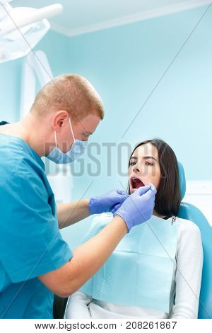 A dermatologist, a blonde man in purple gloves, examines the teeth of a young pretty black-haired girl at his reception