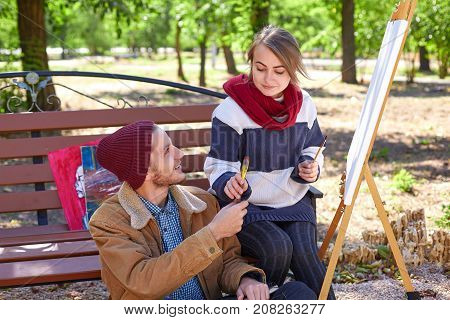 A guy with a beard in a red hat looks like a beautiful blonde girl paints a painter