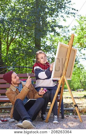 A guy with a beard in a red hat photographes how a beautiful blonde girl paints a picture