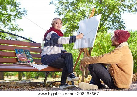 A guy with a beard in a red hat looks like a beautiful blonde girl paints a painter