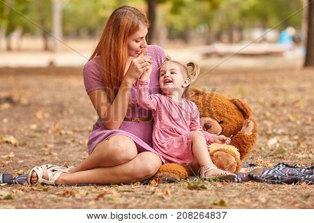 Gorgeous young mother playing with adorable little daughter with a huge plush teddy bear on a blurred autumn park background.