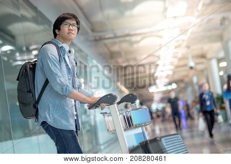 Young Asian man walking with airport trolley and his suitcase luggage in the international airport terminal arrival from travel abroad