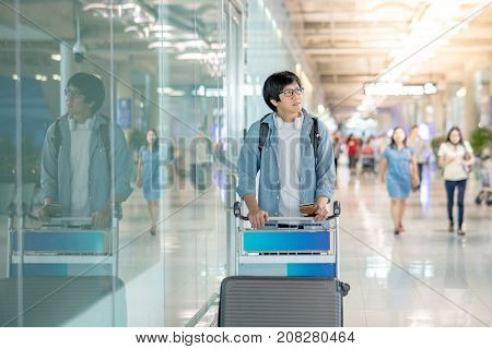 Young Asian man walking with airport trolley and his suitcase luggage in the international airport terminal arrival from travel abroad