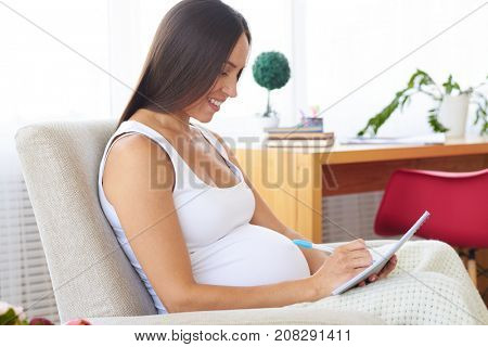 Side view of smiling pregnant woman covered with a blanket writing in jotter while sitting in armchair 