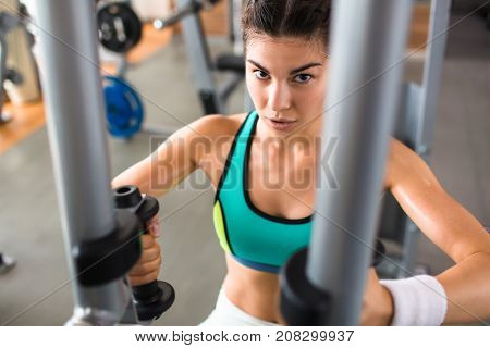Confident athletic woman looking at camera while having intensive training on butterfly machine at modern gym, waist-up portrait