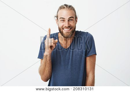 Happy handsome bearded guy with stylish hairstyle brightful smiling with teeth and pointing upside with finger on white background. Copy space.