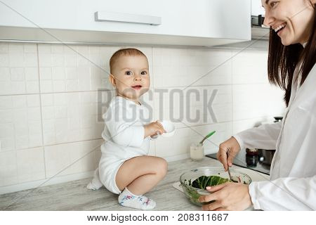 Portrait of cheerful little baby boy sitting on kitchen table, smiling and trying to help mother with cooking salad while mom brightfully smiling