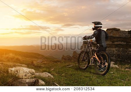 Ryder in full protective equipment on the mtb of two pendant bicycles rides along the green grass towards the sunset in the rays of the sunset sun against the backdrop of a landscape of rocky terrain under the setting sun and clouds.