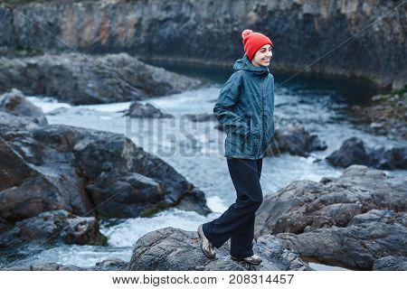 a woman tourist in waterproof clothes and red hat stands on the Bank of the mountain river with small waterfall. Glanni waterfall, Iceland