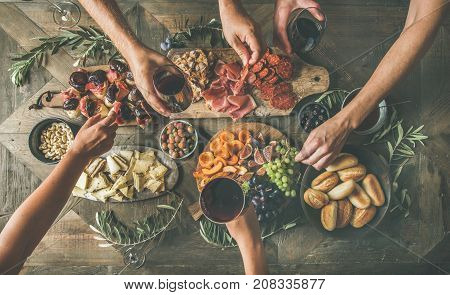 Flat-lay of friends eating and drinking together. Top view of people having party, gathering, celebrating at wooden rustic table set with various wine snacks and fingerfoods. Hands holding glasses