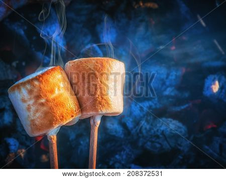 Two brown sweet marshmallows roasting over fire flames. Marshmallow on skewers roasted on charcoals.