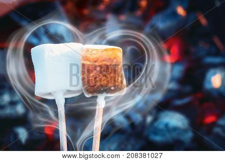 Two white sweet marshmallows roasting over fire flames. Smoke in form of heart. Marshmallow on skewers roasted on charcoals. Sweet love concept
