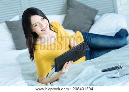 Taking a break. Cheerful young lady looking into the camera with a cheerful smile on her face while relaxing on her bed with a tablet computer in hands.