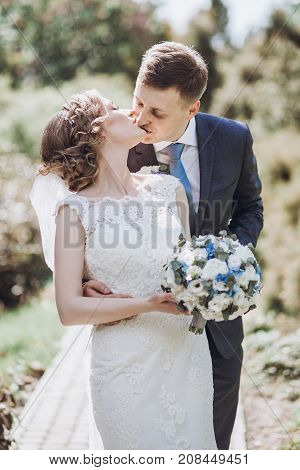 Beautiful Gorgeous Bride And Groom Kissing In Sunny Spring Park And Enjoying Day. Happy Wedding Coup