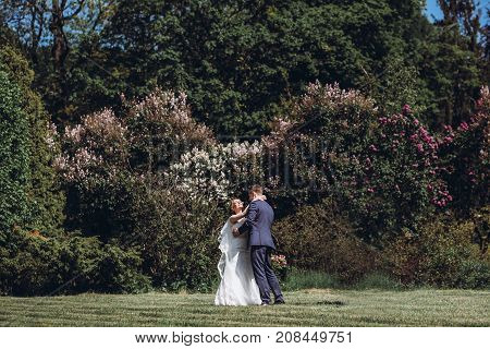 Beautiful Gorgeous Bride And Groom Posing In Sunny Spring Park Among Green Leaves. Happy Wedding Cou