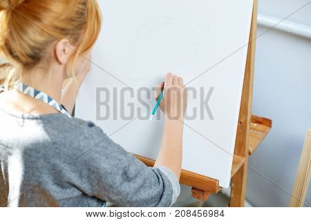 A beautiful artist girl paints on a white canvas with bright multicolored oil paints in a working room. Woman with blonde hair in a blue apron likes to draw. Close-up of hands.