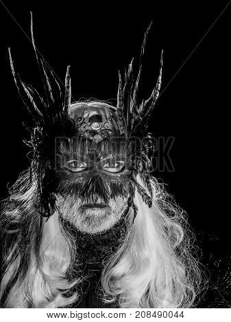 Male bearded face with long white hair and ominous sight in vintage decorative masque with dark brown feathers for fancy ball on black background studio