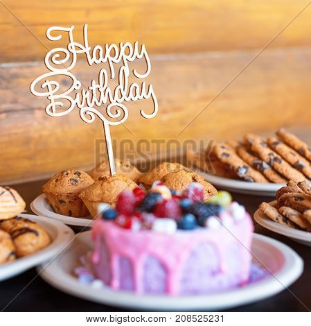 Birthday cake and muffins with wooden greeting sign on rustic background. Wooden sing with letters Happy Birthday and holiday sweets