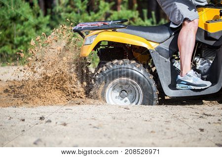 An ATV quadbike get stuck in a sandy road near forest and having wheel-spin making a spray of sand.