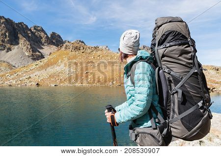 Hiker girl in sunglasses down jacket and hat with a backpack and mountain equipment with tracking sticks in her hands looks at the beautiful view of a high mountain lake in an archipelago in the northern Caucasus. Karachay-Cherkess Republic. 