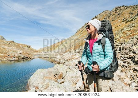 Hiker girl in sunglasses down jacket and hat with a backpack and mountain equipment with tracking sticks in her hands looks at the beautiful view of a high mountain lake in an archipelago in the northern Caucasus. Karachay-Cherkess Republic. 