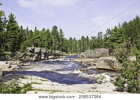 Vertical of rapids running between a rocky shoreline with a rock face in the foreground and surrounded by trees and greenery at Pabineau Falls near Bathurst, New Brunswick on a bright sunny day with blue skies and clouds in August.