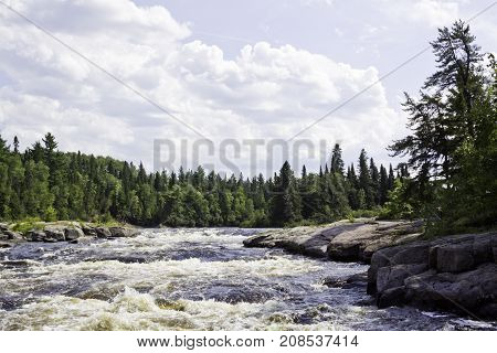 Wide view of rapids running between a rocky shoreline and surrounded by trees and greenery at Pabineau Falls near Bathurst, New Brunswick on a bright sunny day with blue skies and clouds in August.