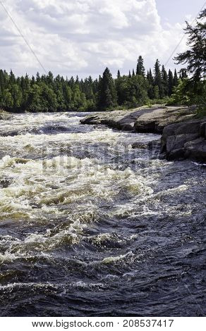 Vertical of rapids running between a rocky shoreline and surrounded by trees and greenery at Pabineau Falls near Bathurst, New Brunswick on a bright sunny day with blue skies and clouds in August.