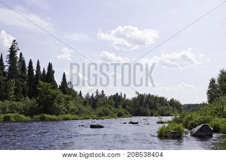 Wide view of a small lake with large rocks on the shoreline and surrounded by trees and greenery at Pabineau Falls near Bathurst, New Brunswick on a bright sunny day with blue skies and clouds in August.