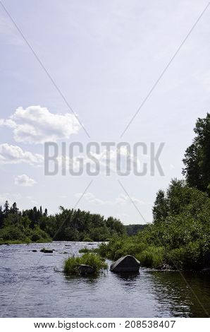 Vertical of a small lake with large rocks on the shoreline and surrounded by trees and greenery at Pabineau Falls near Bathurst, New Brunswick on a bright sunny day with blue skies and clouds in August.