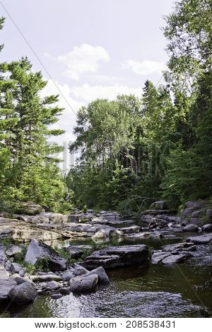 Wide view of a small stream running between boulders and a rocky shoreline surrounded by trees and greenery at Pabineau Falls near Bathurst, New Brunswick on a bright sunny day with blue skies and clouds in August.