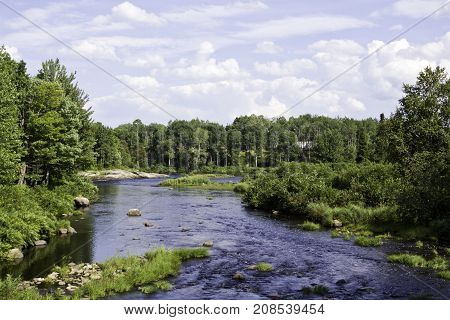 Wide view of a large stream surrounded by trees and greenery at Pabineau Falls near Bathurst, New Brunswick on a bright sunny day with blue skies and clouds in August.
