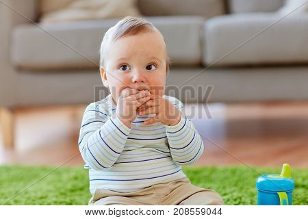 childhood, kids and people concept - baby boy sitting on floor with sippy cup and eating rice cracker at home