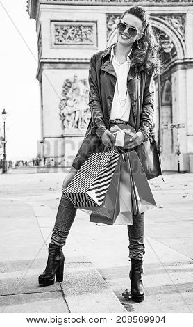 Stylish Christmas in Paris. Full length portrait of happy young woman in sunglasses with shopping bags and Christmas present near Arc de Triomphe in Paris France