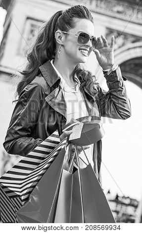 Stylish Christmas in Paris. smiling elegant woman in trench coat with shopping bags and Christmas present near Arc de Triomphe in Paris France looking into the distance