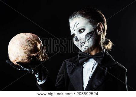 Portrait of a child boy with carnival make-up of a skeleton wearing elegant tail-coat. Studio shot over black background. Halloween. Dia de los muertos. The Day of the Dead.