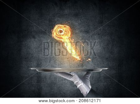 Cropped image of waitress's hand in white glove presenting flaming key symbol on metal tray with dark wall on background.