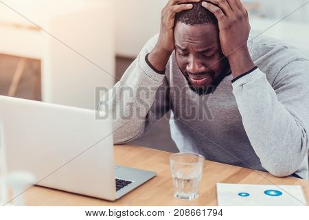 Cannot stand it anymore. Emotionally exhausted young man in casual getting emotional and pressing his head in hands while sitting at a laptop and struggling with a serious project at work.