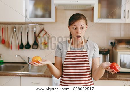 A Young Confused Woman In An Apron Decides To Choose A Red Or Yellow Tomato In The Kitchen. Dieting