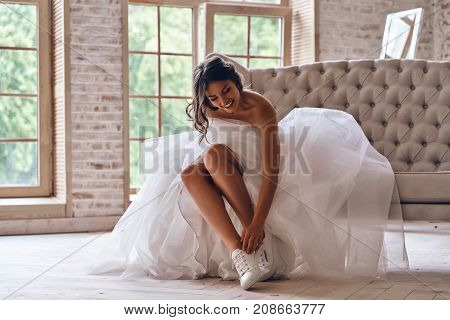 Fits perfectly. Attractive young woman in wedding dress putting on sports shoes and smiling while sitting on the sofa