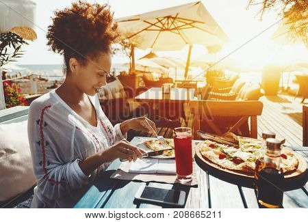 Attractive young black female using knife and spork to cut hot piece of delicious freshly baked pizza with cheese cherry tomatoes and salad while sitting in luxury restaurant outdoor warm sunny day