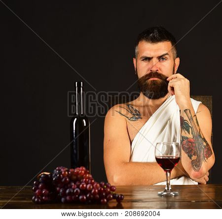 Man with beard near glass of alcohol on brown background. God Bacchus with strict face wearing white cloth sits by wine bottle and grapes. Viticulture and grape harvest concept. Sommelier tasting wine
