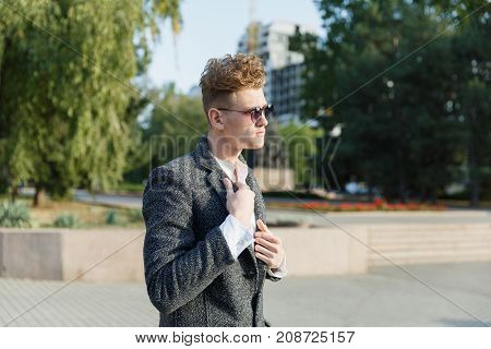 A young guy in the city wearing glasses, against the background of trees in a gray coat. Close-up.